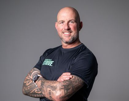 Mark is smiling, with his arms crossed - against a light grey background.