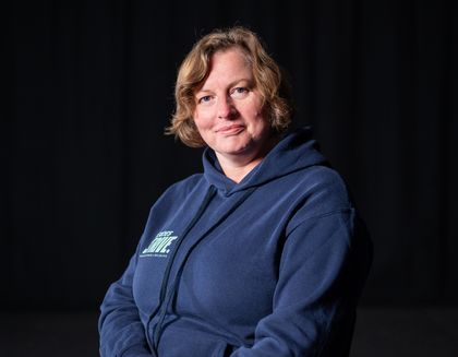 Louise, the yoga instructor is smiling, with her arms crossed - against a dark background.