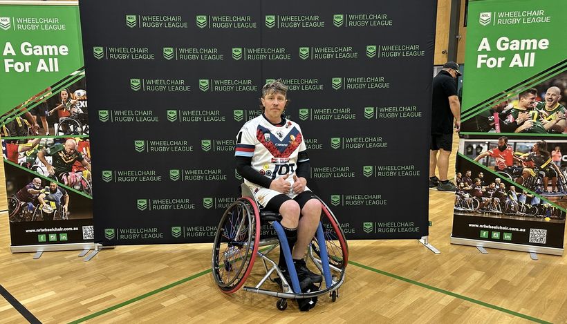 Image shows Ben in n frony of a Wheelchair Rugby League stand