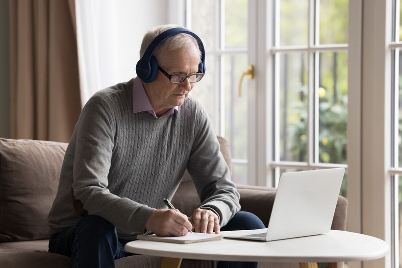 Elderly man writing on a notepad with a laptop in front of him